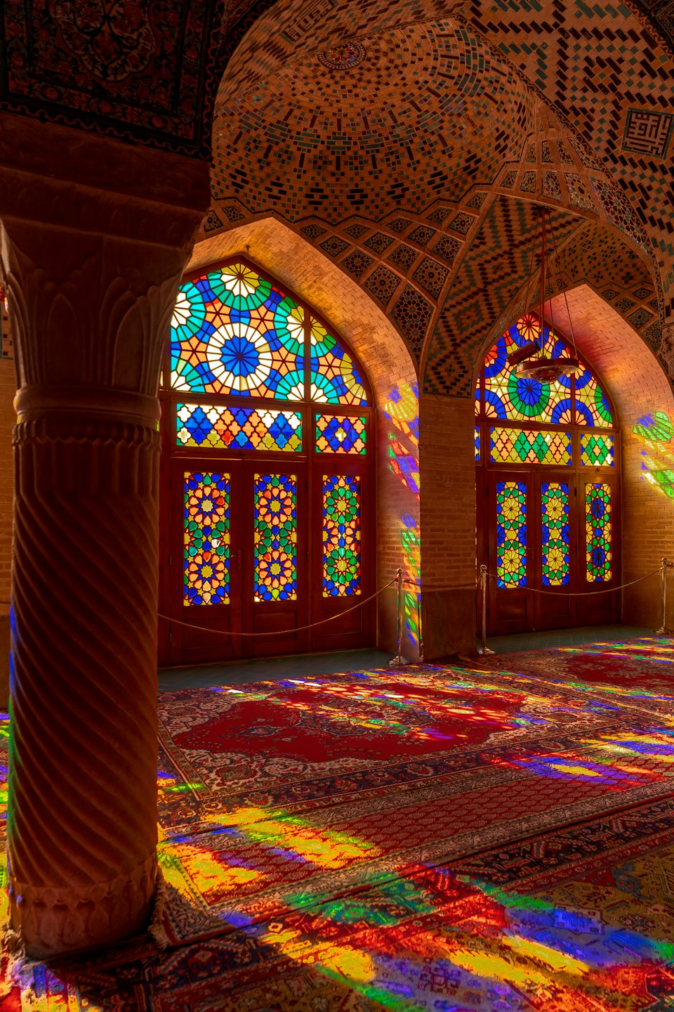 Sunlight streams through arched stained glass windows onto ornate mosque carpets.